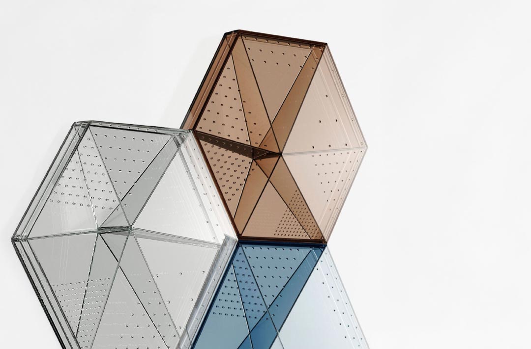 paneles hexagonales de vidrio con tonos transparentes, marrón y azul, con perforaciones distribuidas uniformemente. Los paneles están inclinados y se superponen parcialmente sobre un fondo blanco, creando un efecto geométrico moderno