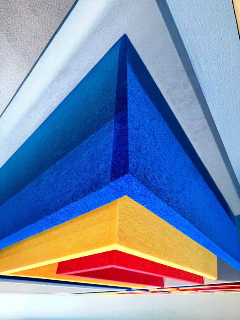 Detalle de un diseño acústico tridimensional con formas geométricas superpuestas en colores vibrantes como azul, amarillo y rojo. Las piezas de material fonoabsorbente crean un efecto visual dinámico y moderno, resaltando su textura y profundidad con una iluminación estratégica