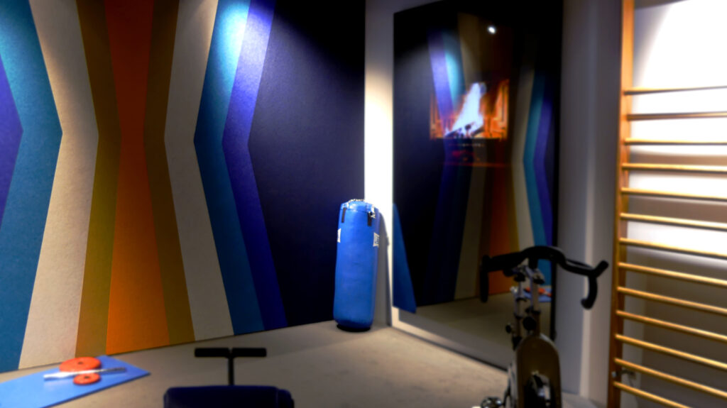 espacio de entrenamiento con paredes decoradas con paneles acústicos en tonos azules, naranjas y blancos, dispuestos en un patrón geométrico. En el centro, un saco de boxeo azul se apoya contra la pared. A la derecha, una bicicleta estática y una espaldera de madera están disponibles para el ejercicio. Un gran espejo refleja la sala, mostrando una pantalla con la imagen de una chimenea encendida. En el suelo, una colchoneta azul con pesas pequeñas y una cuerda de salto completan el área de entrenamiento