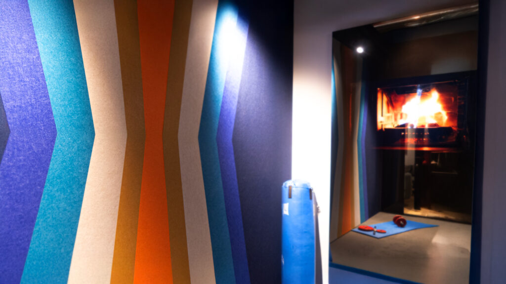 Interior moderno con revestimientos acústicos de colores vibrantes en patrones geométricos. A la derecha, una chimenea con fuego encendido aporta calidez al espacio. En primer plano, un rollo azul y herramientas sugieren un proceso de instalación en curso. Iluminación estratégica resalta la textura y profundidad del diseño