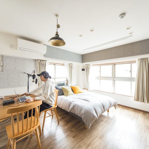 Rectangular-shaped room with soft natural lighting, featuring a wooden desk, minimalistic bed, and acoustic panels for soundproofing. The space includes modern furniture and subtle design elements for an optimal recording environment.